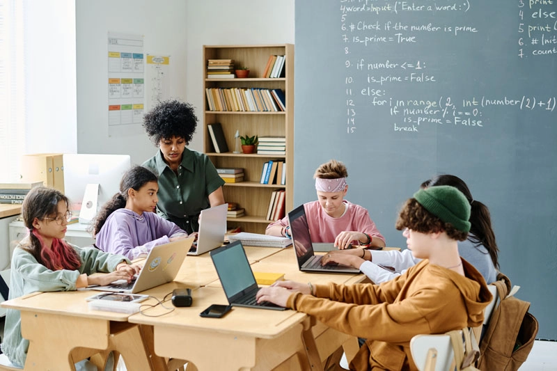 teacher assisting students while using laptops in a classroom setting