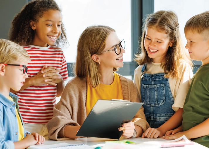 A teacher holding a clipboard smiles at a group of four young students. 