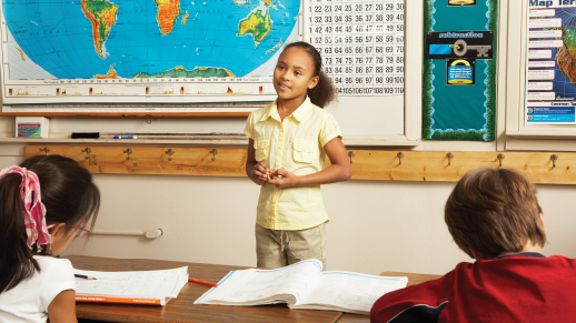 A young student stands at the front of a classroom before a large map of the world. 