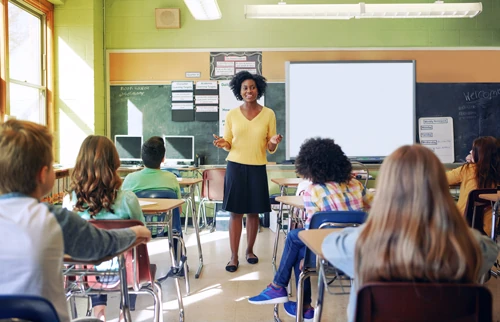 A smiling teacher stands in front of a classroom of students seated at desks.