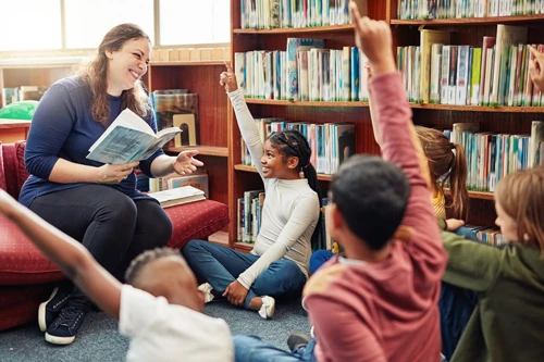 A teacher smiles down at students raising their hands.